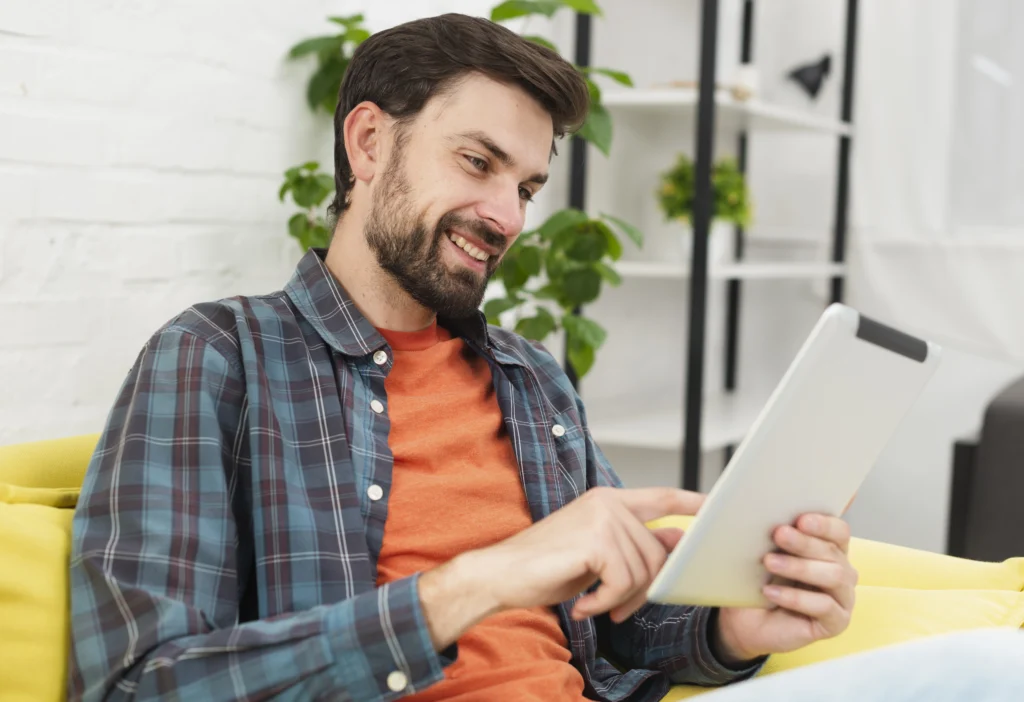 Homem sorrindo enquanto utiliza um tablet no sofá, representando o uso de ferramentas digitais para ABA e acompanhamento terapêutico.